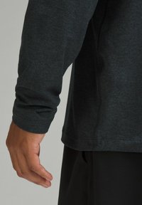Tricou sportiv cu mânecă lungă dintr-un material gri închis. Are un finisaj texturat, cusături plate și un logo pe marginea inferioară.