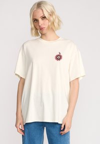 T-shirt oversize couleur crème en coton doux, avec un motif de fleur rose et un œil sur la poitrine, à manches courtes et à coupe décontractée.