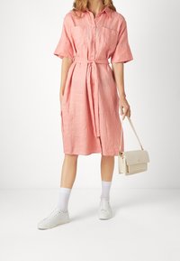 Abito camicia di lino rosa con maniche corte, apertura con bottoni, cintura in vita e due tasche sul petto. Abbinato a sneaker bianche e una borsa leggera.