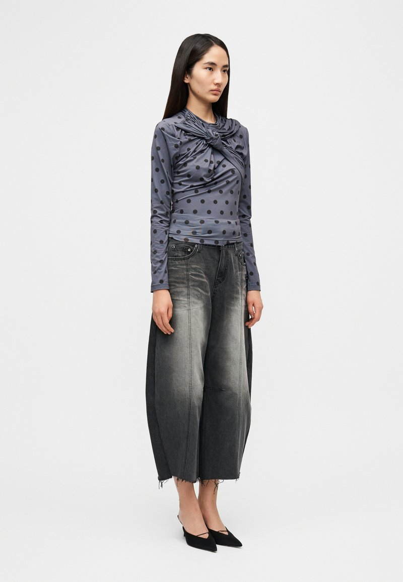 Haut gris à manches longues avec pois noirs et encolure drapée ; pantalon large en denim gris à texture délavée ; chaussures noires à bouts pointus.