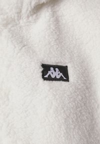 Tela de forro blanco con un pequeño parche negro que presenta un logo blanco de dos figuras sentadas de espaldas una a la otra.