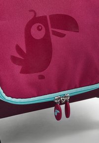 Magenta stoffen tas met een getextureerd papegaaiontwerp. Kenmerkt een turquoise rits en ronde ritstrekker met een kleine blauwe vogelafbeelding.