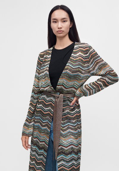 Femme portant un long cardigan multicolore à motif zigzag par-dessus un top noir et un jean, une main sur la hanche devant un fond uni.
