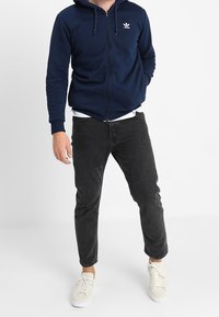 Mörkblå zip-hoodie med huva och liten vit logotyp, ihopparad med svarta jeans och ljusa sneakers. Standard passform, avslappnad stil.