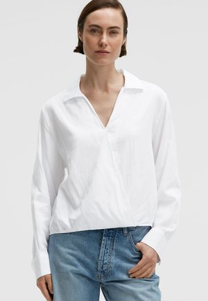 Frau trägt eine weiße Langarmbluse mit tiefem V-Ausschnitt und blaue Jeans, steht mit einer Hand in der Tasche vor einem schlichten Hintergrund.