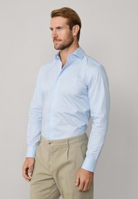 Man met lichtbruin haar, gekleed in een lichtblauwe langarmige overhemd dat in beige broeken is gestoken, staand tegen een effen achtergrond.