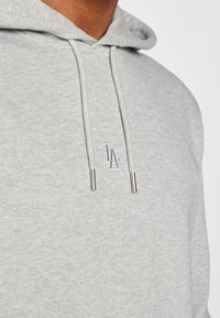 Light gray hoodie featuring a kangaroo pocket, drawstrings with metal tips, and a small embroidered logo on the chest.