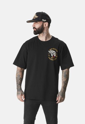 T-shirt en coton noir avec un col rond et des manches courtes. Présente un imprimé graphique doré et blanc sur le côté gauche de la poitrine. Le modèle porte un jean noir et une casquette.