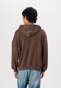 Barna, oversize kapucnis pulóver, texturált felülettel és húzózsinórral. Világoskék farmernadrággal párosítva. Hátsó nézet, amely a laza szabást mutatja.