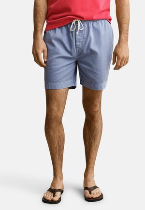 Mann trägt hellblaue Shorts mit Kordelzug, rotes Hemd und schwarze Flip-Flops, steht mit entspannt an den Seiten hängenden Händen.