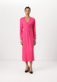 Fuchsia long-sleeve dress with a deep V-neck and knotted front detail. Smooth, flowing fabric, ankle-length, worn with black high heels.