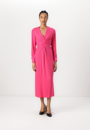 Fuchsia long-sleeve dress with a deep V-neck and knotted front detail. Smooth, flowing fabric, ankle-length, worn with black high heels.