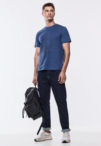 Modré bavlněné tričko s textem v proužkovaném designu, tmavě modré džíny, šedé tenisky a černý batoh. Jednoduchá, neformální kompozice.