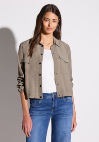 Veste légère vert olive avec des boutons, deux poches avant et des poignets élastiques, portée sur une chemise blanche et un jean bleu.