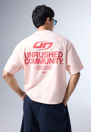 Bărbat cu spatele la cameră purtând un tricou roz pal cu text roșu „UNRUSHED COMMUNITY” și un text mai mic dedesubt, asortat cu pantaloni scurți închiși la culoare.