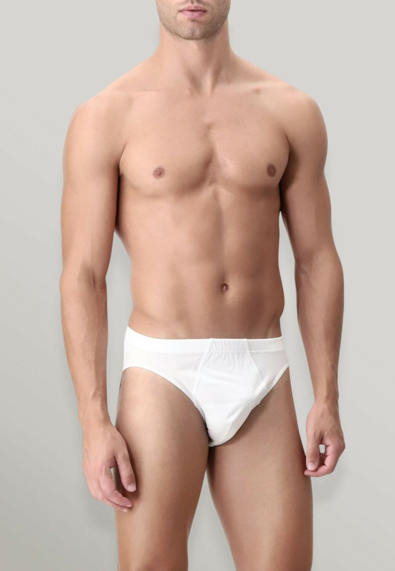 Slip bianco in cotone con elastico in vita, design aderente e texture liscia. Il modello lo mostra su uno sfondo grigio neutro.