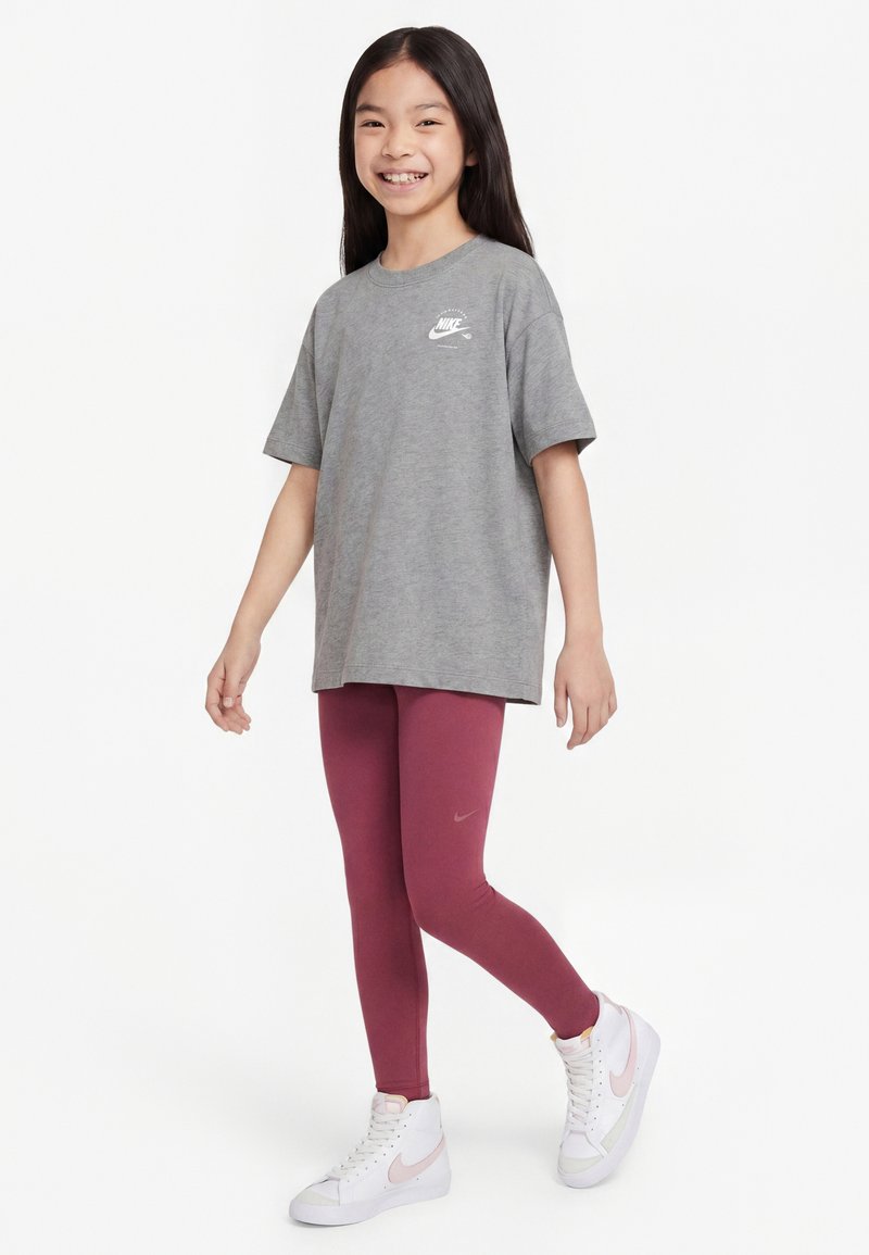Fille souriante debout, portant un t-shirt Nike gris oversized, des leggings bordeaux Nike et des baskets montantes blanches avec un swoosh rose.