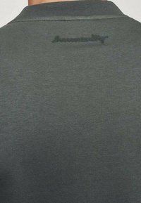 Ciemnoszary materiał z delikatnym wyszytym napisem blisko górnej części środka, pokazujący zbliżenie na tył kołnierza koszuli.