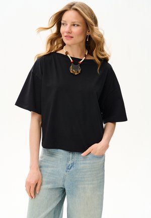 Kobieta o falowanych blond włosach, ubrana w luźną czarną koszulkę z krótkim rękawem, jasnoniebieskie dżinsy i naszyjnik z koralików z dużym wisiorkiem.