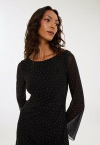 Czarna sukienka z długimi, półprzezroczystymi rękawami, charakteryzująca się dopasowanym krojem i białym wzorem w kropki. Gładka tekstura z delikatnym połyskiem.