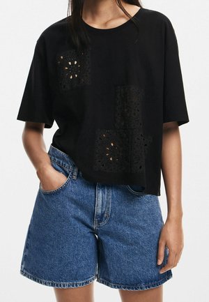 Person trägt ein locker sitzendes schwarzes T-Shirt mit gemusterten Ausschnitten und blaue, hoch taillierte Jeansshorts, steht mit einer Hand in der Tasche.