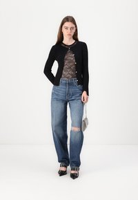 Cardigan nero sopra un top in pizzo, jeans chiari strappati, tacchi neri a punta con borchie e una piccola borsa metallica, in posa su uno sfondo bianco.