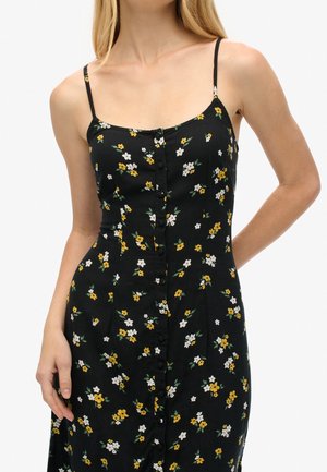 Femme portant une robe noire sans manches avec de petits motifs floraux jaunes et blancs et des boutons sur le devant, debout avec une main derrière le dos.