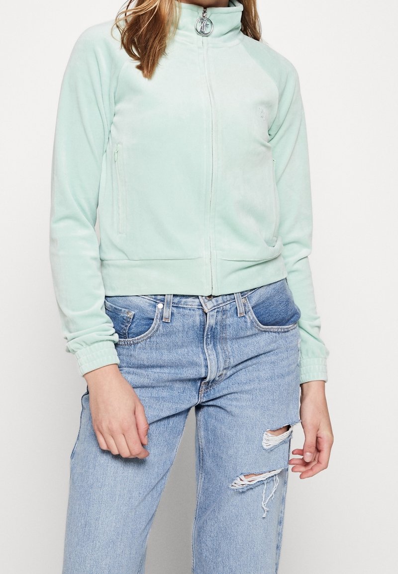 Personne portant une veste zippée vert menthe clair et un jean bleu taille haute avec une déchirure à un genou, debout devant un fond blanc uni.