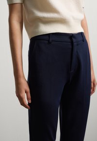 Marineblauwe broek met een getailleerde pasvorm, gemaakt van een gladde stof, voorzien van voorplooien en zijzakken, gecombineerd met een lichtbeige top.