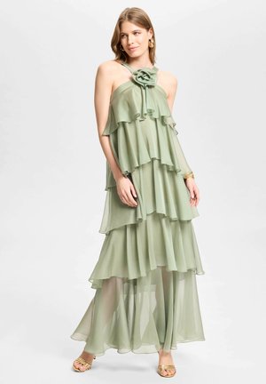 Femme portant une robe maxi à volants vert clair avec un détail floral au niveau de l'encolure, associée à des sandales dorées et des boucles d'oreilles créoles.