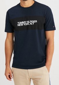 Mand iført en navy blue Tommy Hilfiger T-shirt med hvid logo-tekst og beige bukser, stående med den ene hånd i lommen.
