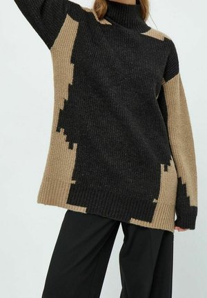 Femme portant un pull à col roulé oversized en maille avec des blocs de couleur abstraits en noir et beige, associé à un pantalon noir ample.