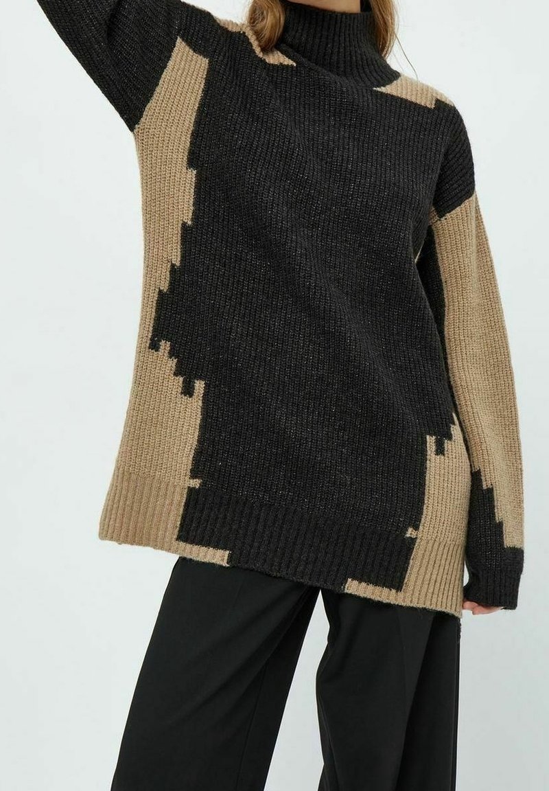 Femme portant un pull à col roulé oversized en maille avec des blocs de couleur abstraits en noir et beige, associé à un pantalon noir ample.