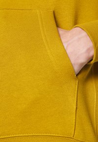 Hoodie amarelo com uma textura suave, apresentando um bolso frontal e punhos canelados. O detalhe das costuras realça o design geral.