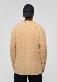 Beige kabelgebreide trui met lange mouwen, een ronde halslijn en een zachte, pluizige textuur. Gedragen met een zwarte pet met witte tekst.