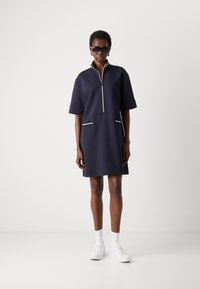 Navy short-sleeve dress with a front zip closure, two front pockets, and white piping details; worn with white sneakers and ankle socks.