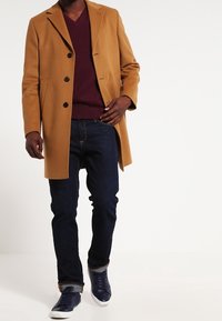 Homme portant un manteau camel, un pull bordeaux, un jean bleu foncé et des baskets bleu marine, marchant sur un fond blanc uni.