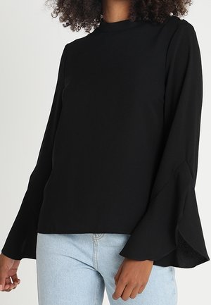 Schwarze langärmlige Bluse mit ausgestellten Manschetten und Perlenknopfdetail an der Schulter, getragen mit hellblauen Jeans.