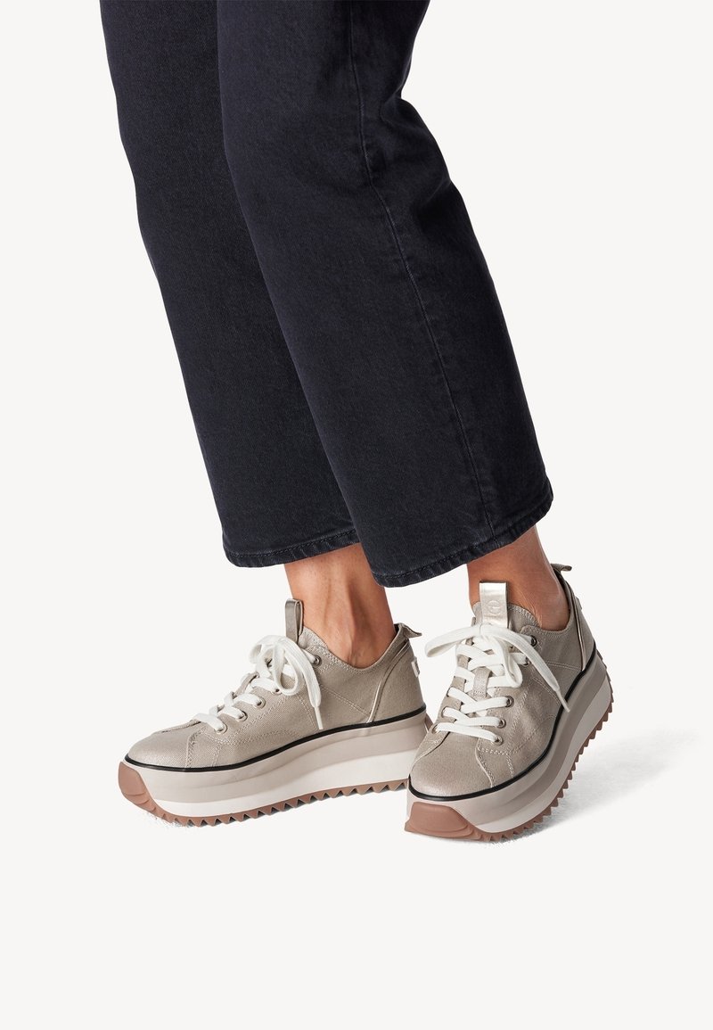 Baskets plateformes beiges claires en matériau texturé, avec des lacets blancs, une bordure noire et une semelle extérieure tan avec un relief visible, associées à un pantalon sombre.
