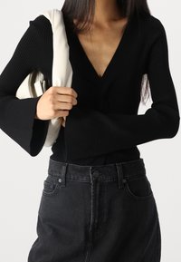 Black ribbed long-sleeve top with a deep V-neck and flared cuffs, paired with high-waisted black denim jeans. A cream shoulder bag is held.