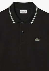 Svart pikétröja med en kontrasterande krage i grön och vit rand, två knappslå, och en broderad grön krokodil-logotyp.