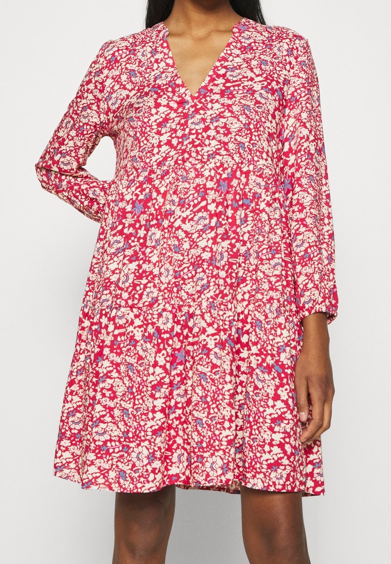 Femme portant une robe rouge à fleurs avec un col en V et des manches longues, debout avec une main sur la hanche devant un fond uni.