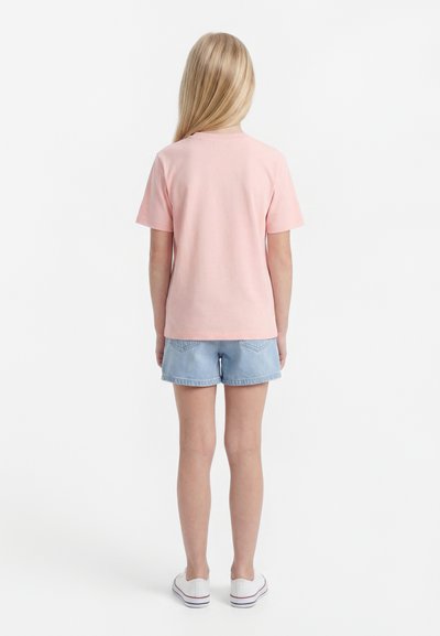 Enfant aux longs cheveux blonds portant un t-shirt rose à manches courtes, un short en denim bleu clair et des baskets blanches, debout de dos.