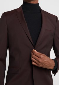 Donker kastanjebruin colbert met notch-lapels, een enkele borstzak en twee voorzakken, gedragen over een zwart geribbelde coltrui.
