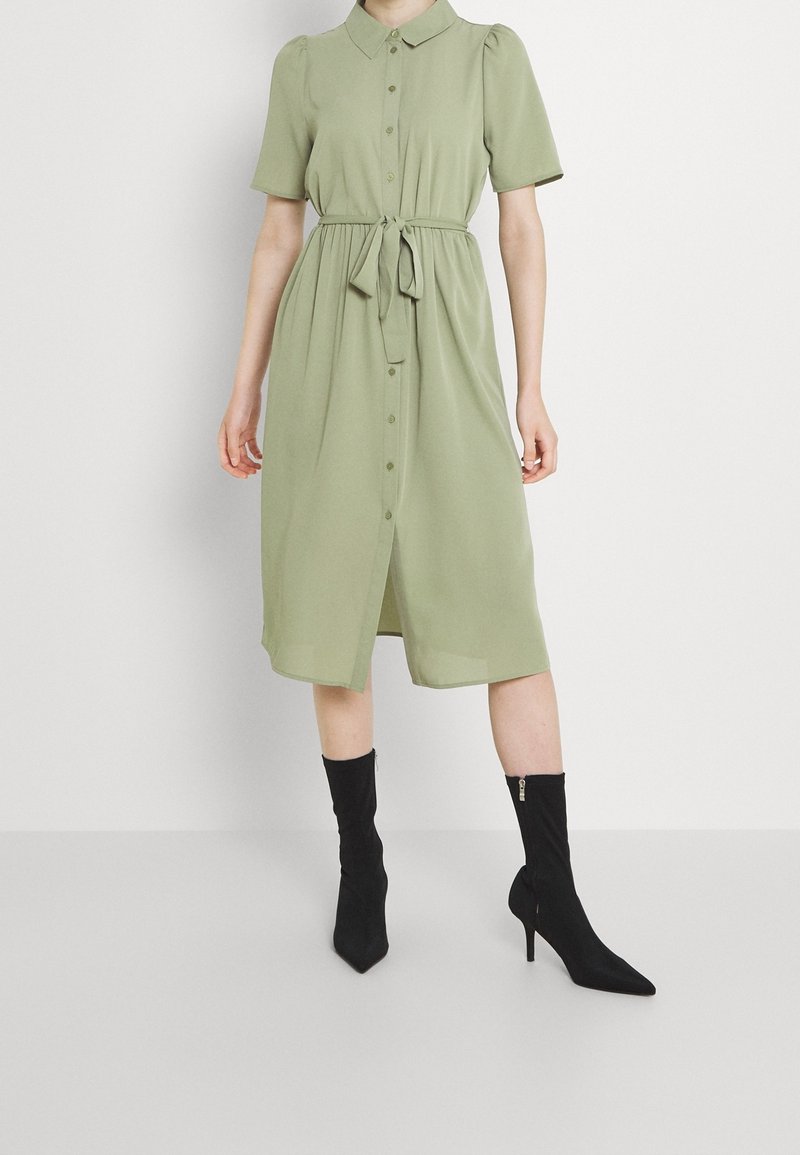 Personne portant une robe boutonnée vert clair à manches courtes avec une taille nouée, associée à des bottines noires à talons.