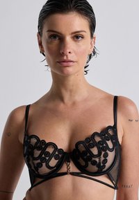 Reggiseno in pizzo nero con motivi floreali, texture in rete, supporto con ferretto e spalline regolabili con chiusura frontale.