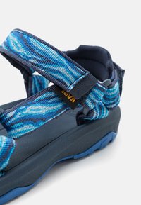 Teva sandal med en mørkeblå gummisåle, med mønstrede blå stropper laget av vevd stoff og justerbare lukkinger for tilpasning.