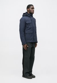 Granatowa ocieplana kurtka z kapturem, wieloma kieszeniami z przodu oraz zamkiem błyskawicznym. Noszona z ciemnymi spodniami i czarnymi trampkami.