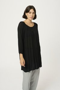 Schwarze, langärmlige Tunika mit Rundhalsausschnitt, einer Knopfdetaillierung an der Vorderseite und plissierter Textur; kombiniert mit hellgrauen Hosen.