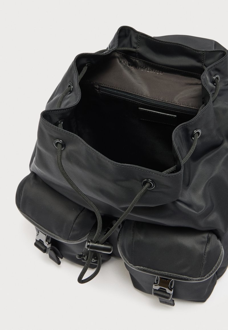 Zaino nero con coulisse con due tasche anteriori con fibbia e un vano principale aperto che mostra una tasca interna con cerniera.